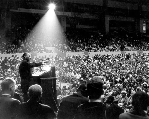 Martin Luther King speaking to a large crowd, standing at a podium, with a beam of light shining down on him.